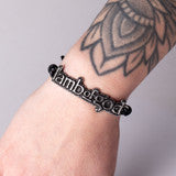 Close-up of a Lamb of God bracelet with the band’s sculpted logo on a black waxed cord, adjustable up to 10 inches in diameter, set against a dark background.